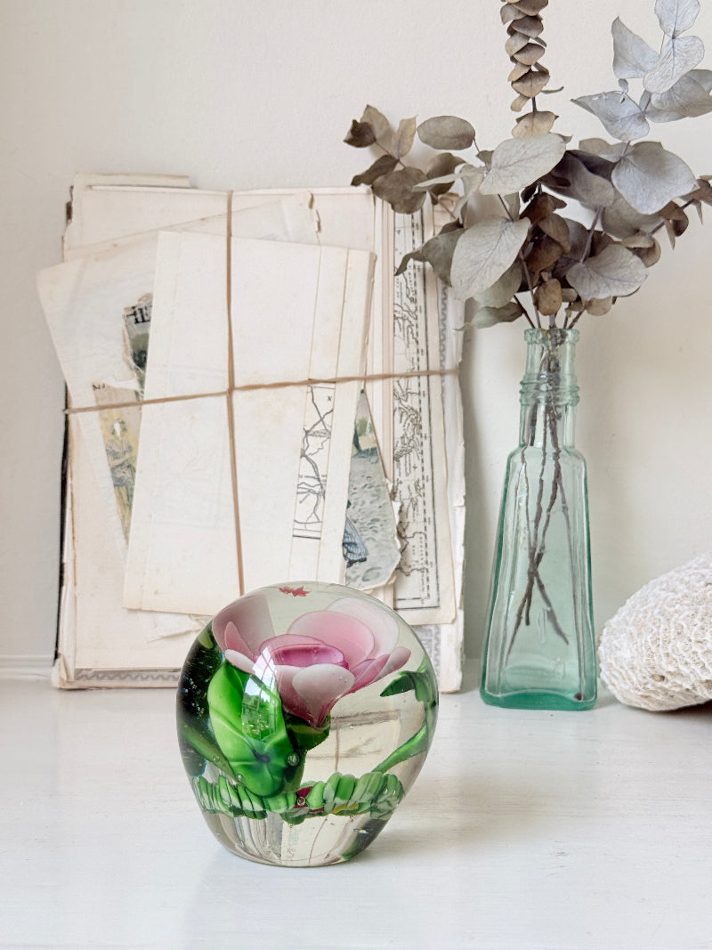 Large clear glass paperweight with a pink flower and green leaves inside, photographed on a white surface