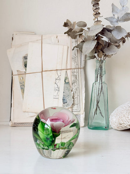 Large clear glass paperweight with a pink flower and green leaves inside, photographed on a white surface