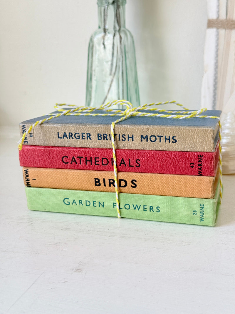 A stack of four vintage hardcover books tied with yellow-and-white string, placed on a white surface with a blurred glass bottle in the background. The book spines read &
