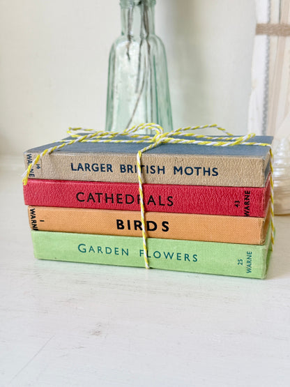 A stack of four vintage hardcover books tied with yellow-and-white string, placed on a white surface with a blurred glass bottle in the background. The book spines read &
