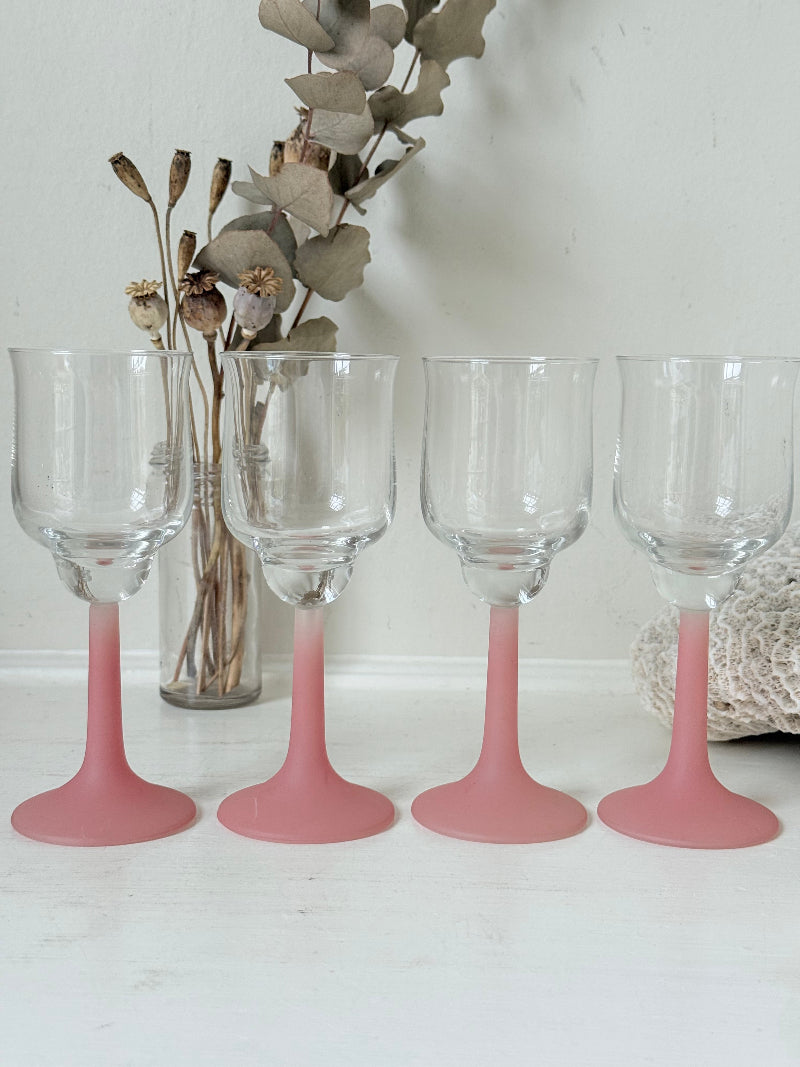 Set of four small vintage wine glasses with clear bowls and pink stems photographed on a white surface.