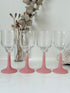 Set of four small vintage wine glasses with clear bowls and pink stems photographed on a white surface.