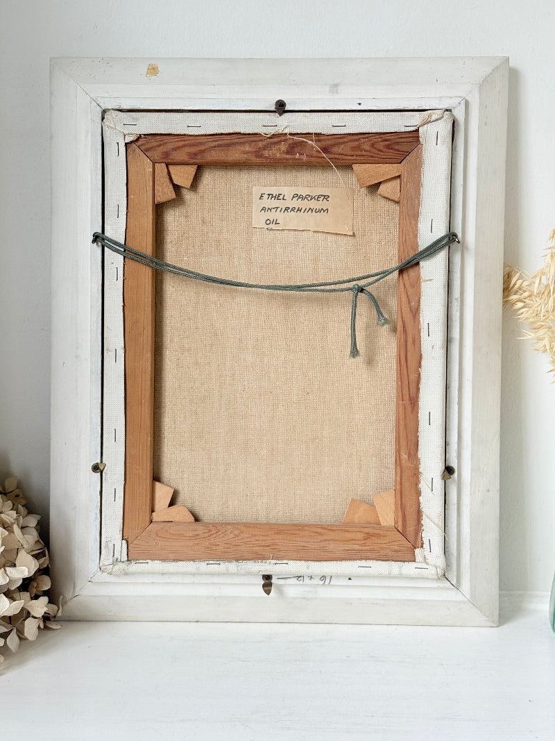 The back view of a framed art print, showing the canvas, wooden stretcher bars, hanging cord, and a handwritten label card, set against a white wall with dried floral decor.