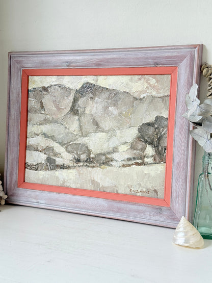 A framed landscape art print featuring textured gray and beige mountains, styled on a white tabletop with a rustic distressed frame, coral inner border, and decorative props.