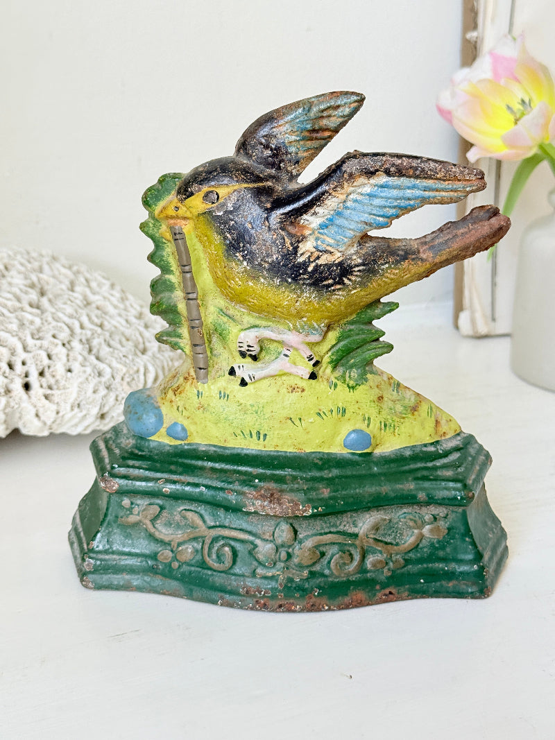 Vintage hand-painted metal bird figurine perched on a green base, holding a pipe, displayed on a white surface with a lace doily and flowers in the background.