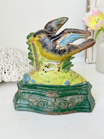 Vintage hand-painted metal bird figurine perched on a green base, holding a pipe, displayed on a white surface with a lace doily and flowers in the background.