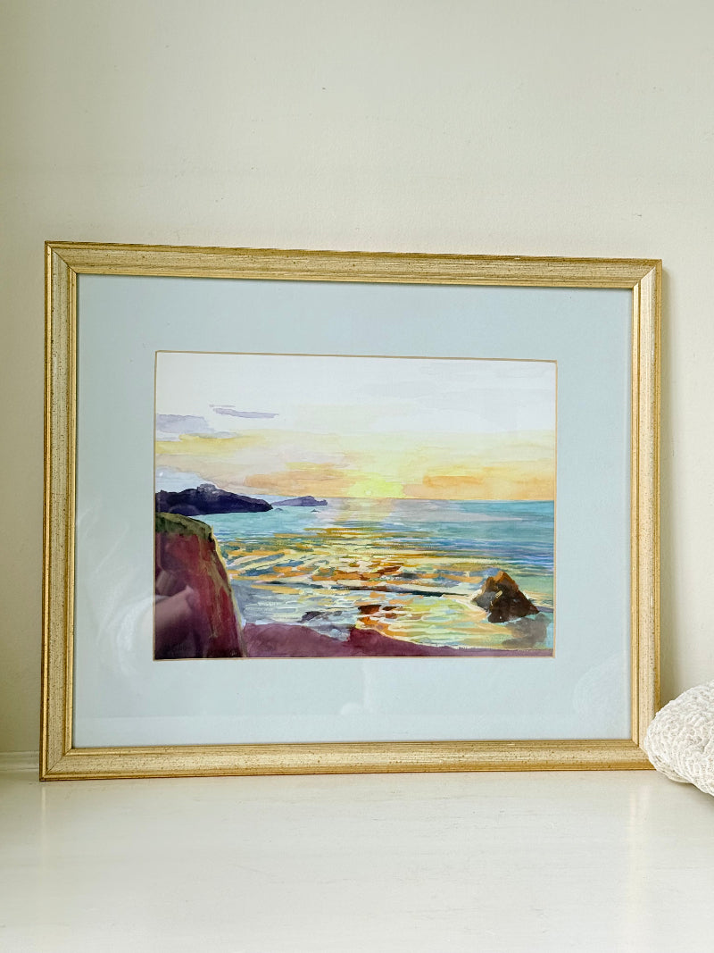 Framed coastal watercolour of sunset over sea with rocks in foreground, in gilt frame with pale mount, photographed on white surface.