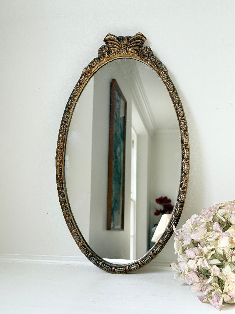 Vintage oval wall mirror with decorative gilt frame and bow crest, styled against a neutral wall with hydrangeas.