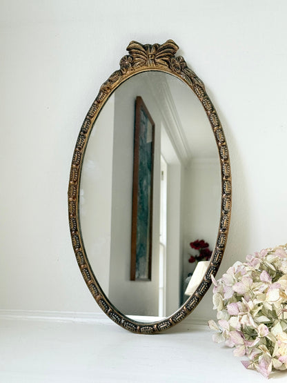 Vintage oval wall mirror with decorative gilt frame and bow crest, styled against a neutral wall with hydrangeas.
