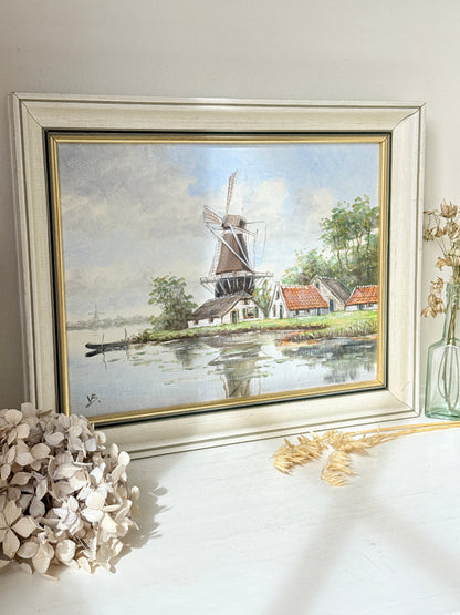 A framed landscape painting of a Dutch windmill by a river, displayed on a white surface with dried flowers and a glass vase nearby. The painting features a light blue sky, green trees, and a brown windmill, enclosed in a distressed white frame with gold inner trim.