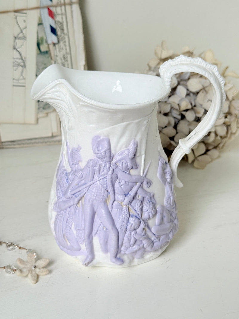 A white ceramic pitcher with a pale lavender embossed classical scene on the front, styled on a tabletop with dried flowers and framed art in the background.