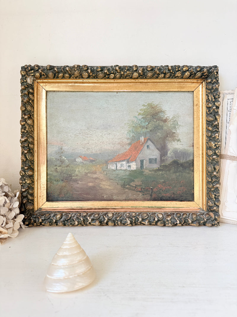 A framed vintage landscape painting depicting a small white house with a red roof on a dirt path, surrounded by trees. The painting is housed in an ornate, textured dark frame with a gold inner border, displayed on a white surface with a seashell and dried flowers as props.