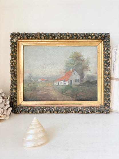 A framed vintage landscape painting depicting a small white house with a red roof on a dirt path, surrounded by trees. The painting is housed in an ornate, textured dark frame with a gold inner border, displayed on a white surface with a seashell and dried flowers as props.