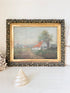 A framed vintage landscape painting depicting a small white house with a red roof on a dirt path, surrounded by trees. The painting is housed in an ornate, textured dark frame with a gold inner border, displayed on a white surface with a seashell and dried flowers as props.
