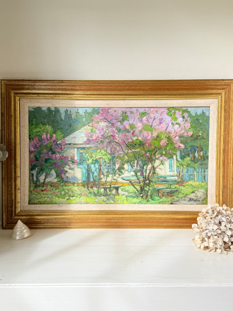 A framed landscape oil painting of a cottage garden with pink flowering trees, displayed on a white shelf with decorative seashells against a neutral wall.