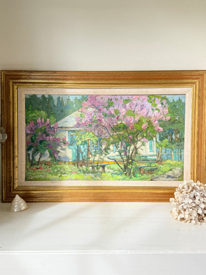 A framed landscape oil painting of a cottage garden with pink flowering trees, displayed on a white shelf with decorative seashells against a neutral wall.