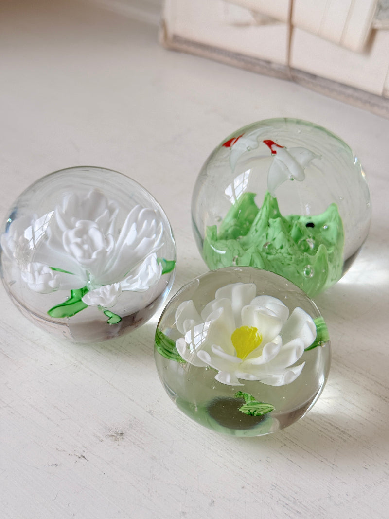 Trio of white flower glass paperweights in graduated sizes with green leaves, photographed on a white surface