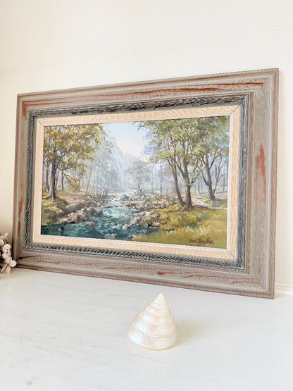 A framed landscape painting of a forest stream, displayed on a white surface against a white wall. The rustic frame has a weathered gray and brown finish, and a small white seashell sits in front of the frame.