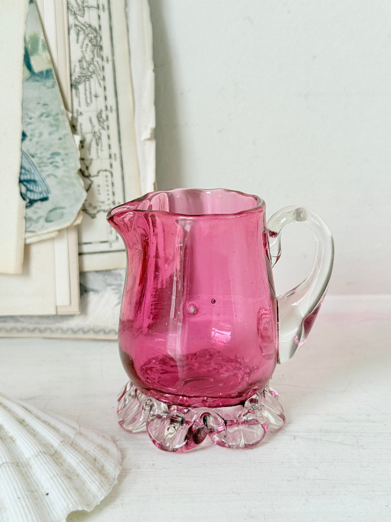 Antique cranberry glass cream jug with clear applied handle, crimped pedestal foot and deep pink hand blown glass body