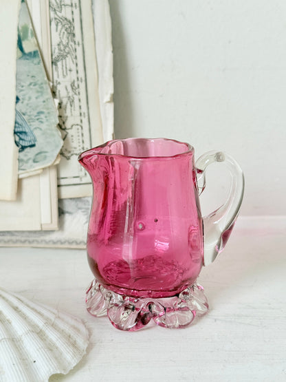 Antique cranberry glass cream jug with clear applied handle, crimped pedestal foot and deep pink hand blown glass body