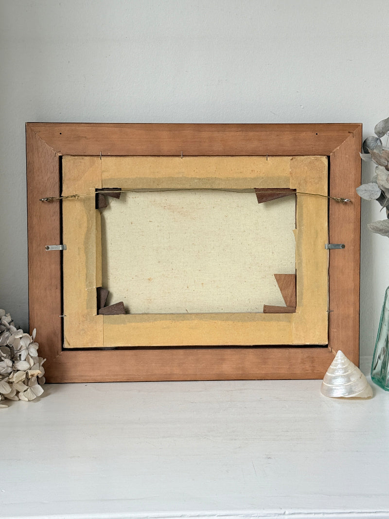 The back of a vintage wooden picture frame, showing the canvas backing, mounting hardware, and tape, styled on a white surface with decorative seashells and dried flowers.