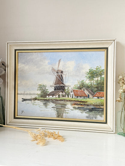 A framed landscape painting of a Dutch windmill by a canal, displayed on a white surface against a light beige wall. The artwork features a windmill, small houses with red-tiled roofs, green trees, and reflections in the water, enclosed in a wide white frame with gold inner trim.
