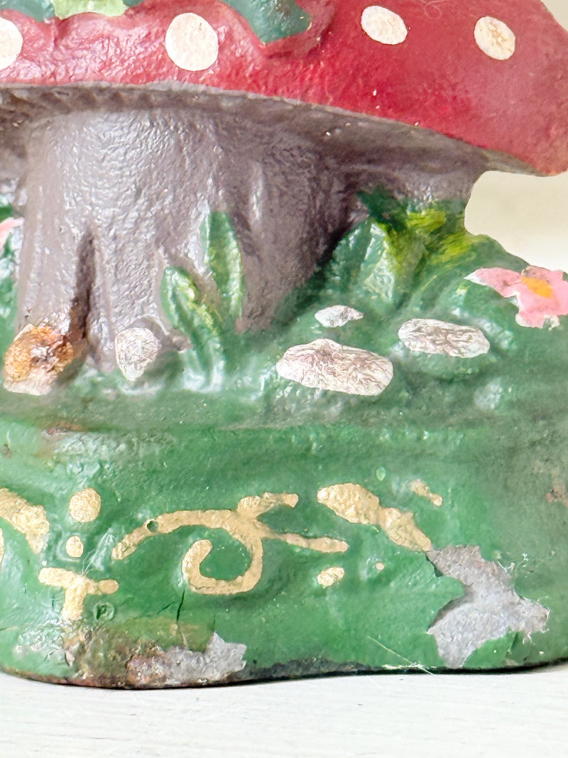 A close-up of a hand-painted cast iron mushroom planter. The design features a red cap with white spots, a dark brown textured stem, and a green base decorated with gold leaf accents, white stones, and a small pink flower.