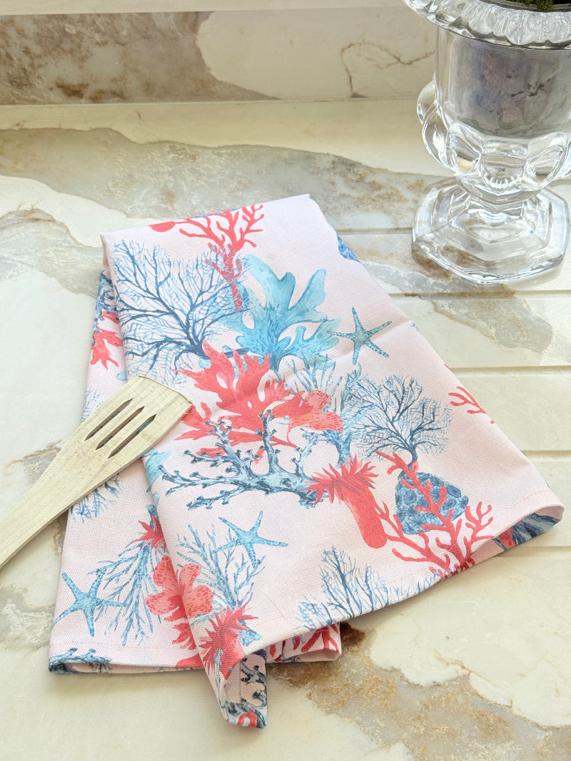Pale pink coral and seaweed print cotton tea towel with wooden spoon on a marble surface beside cut glass