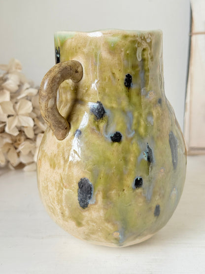 A handmade ceramic pitcher with a rounded body, a rustic loop handle, and a glossy glaze featuring olive green, beige, and black speckles, set against a softly blurred background.
