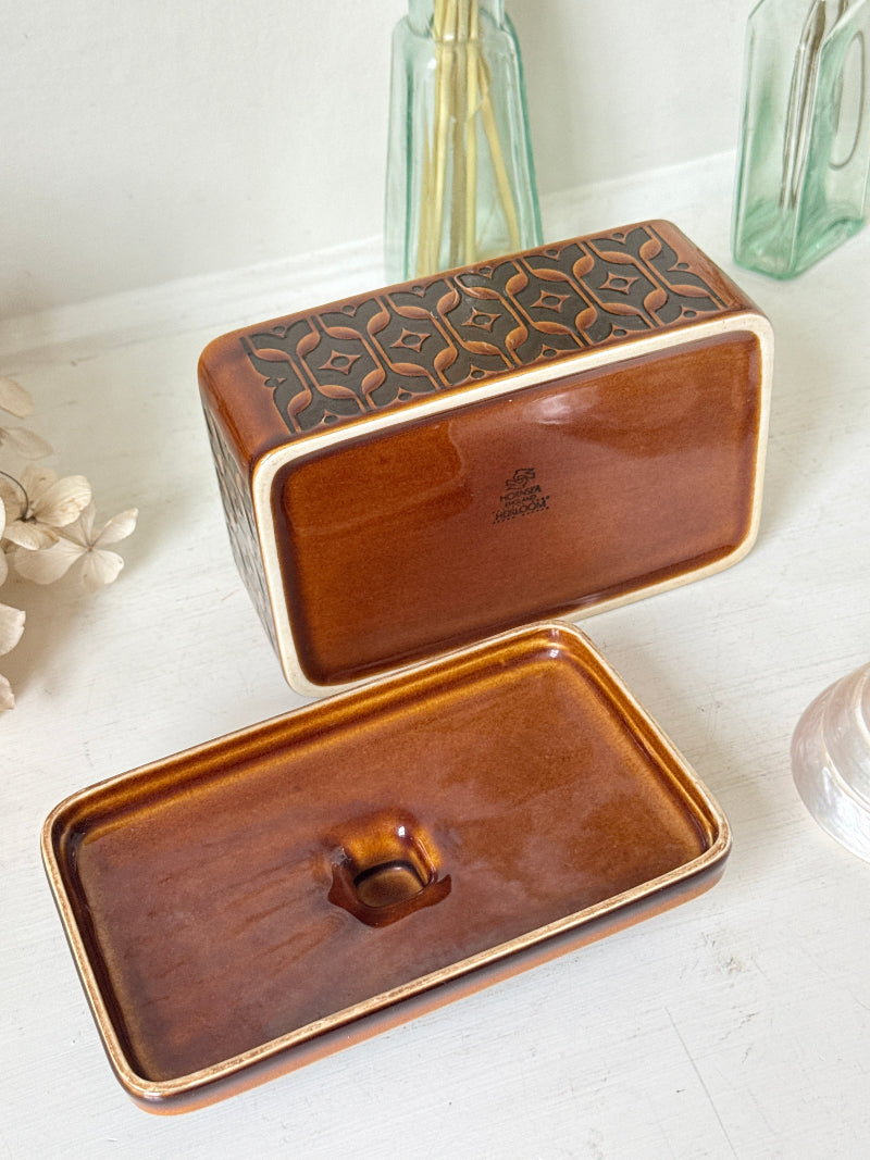 A rectangular ceramic soap dish with a glossy amber-brown glaze and a raised geometric pattern on the lid, shown open with the dish placed below. The items are styled on a white surface with soft-focus decorative glass bottles in the background.