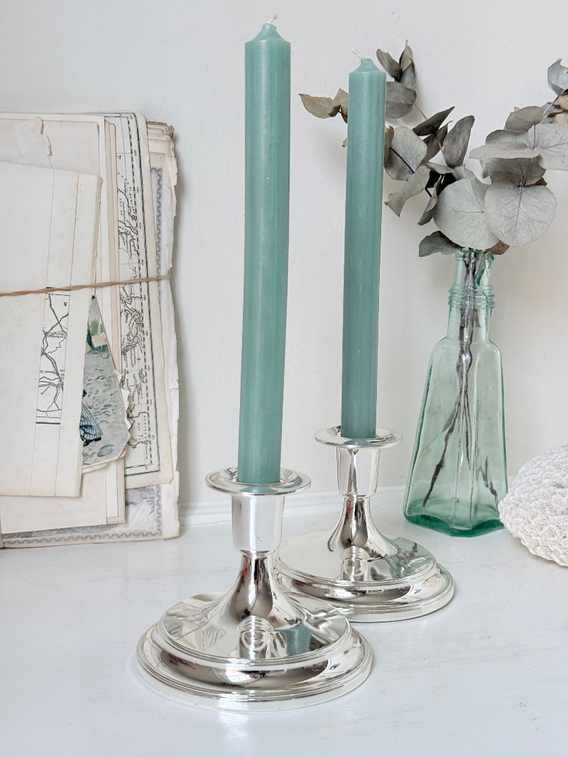 Pair of silver tone candlesticks with wide round stepped bases holding green candles, photographed on a white surface