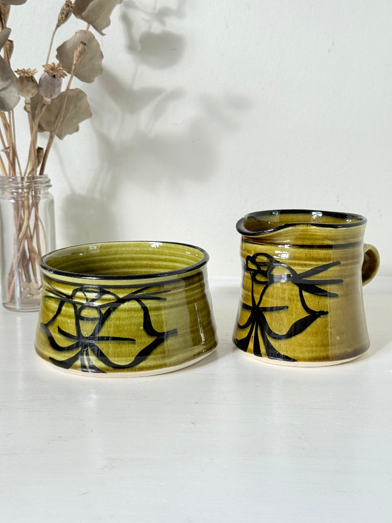 Studio pottery sugar bowl and milk jug in olive green glaze with black brushstroke decoration, photographed on a white surface.