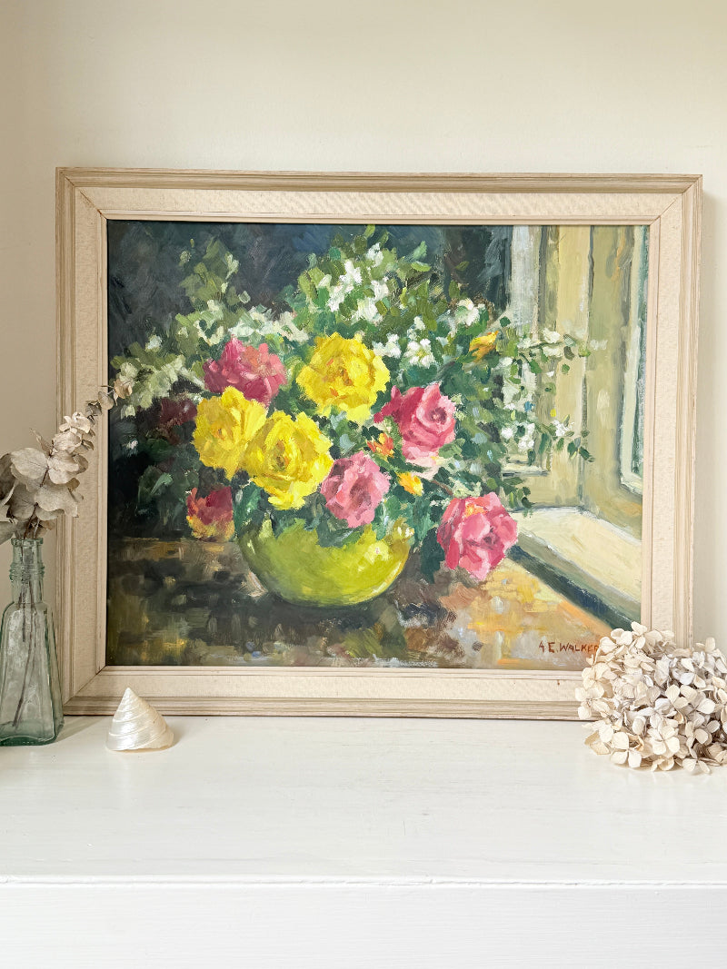 A framed still life oil painting of a vibrant flower bouquet in a green vase, displayed on a white mantelpiece or shelf with decorative seashells and a glass vase against a beige wall.