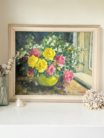 A framed still life oil painting of a vibrant flower bouquet in a green vase, displayed on a white mantelpiece or shelf with decorative seashells and a glass vase against a beige wall.