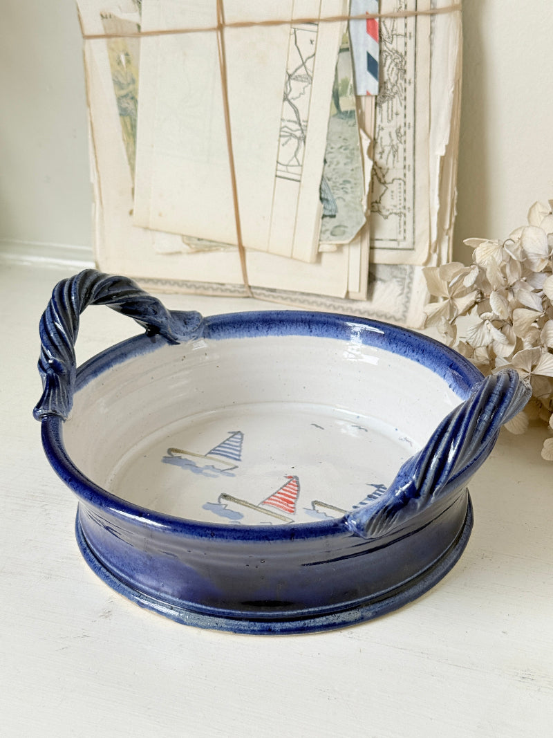 A round ceramic bowl with twisted side handles, featuring a glossy navy blue exterior and a white interior decorated with small sailboat illustrations. The bowl is styled on a light surface with vintage papers and dried flowers in the background.