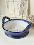 A round ceramic bowl with twisted side handles, featuring a glossy navy blue exterior and a white interior decorated with small sailboat illustrations. The bowl is styled on a light surface with vintage papers and dried flowers in the background.