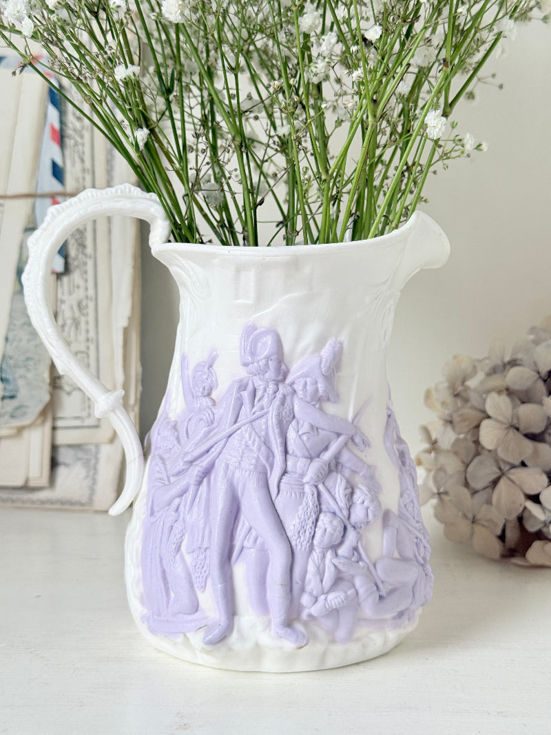 A white ceramic pitcher vase with pale lavender embossed classical figures, filled with baby&