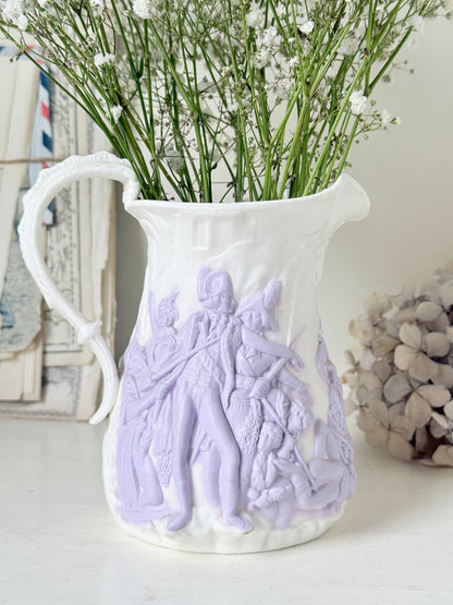 A white ceramic pitcher vase with pale lavender embossed classical figures, filled with baby&