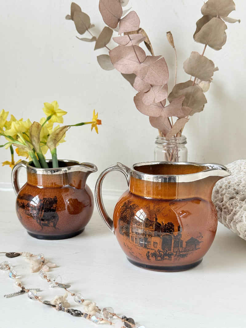 Pair of antique Ridgways stoneware creamer jugs photographed on a white surface in amber treacle glaze with black coaching scenes and silver lustre rims
