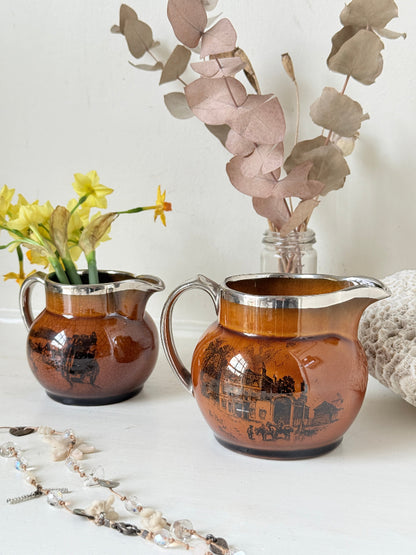 Pair of antique Ridgways stoneware creamer jugs photographed on a white surface in amber treacle glaze with black coaching scenes and silver lustre rims
