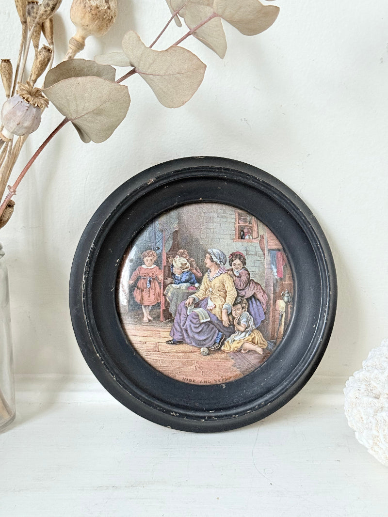 Antique Prattware pot lid in black round frame with Victorian family scene on white surface.