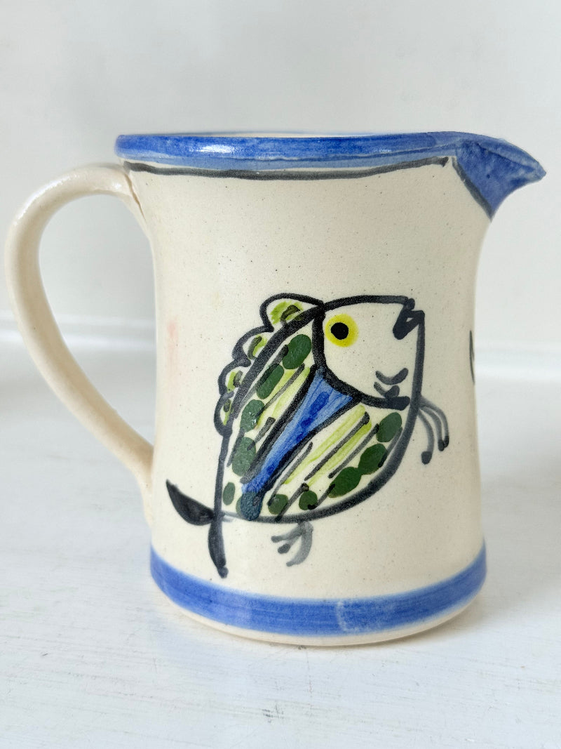 Vintage studio pottery jug with hand painted blue and green fish design on cream clay, photographed on a white surface.
