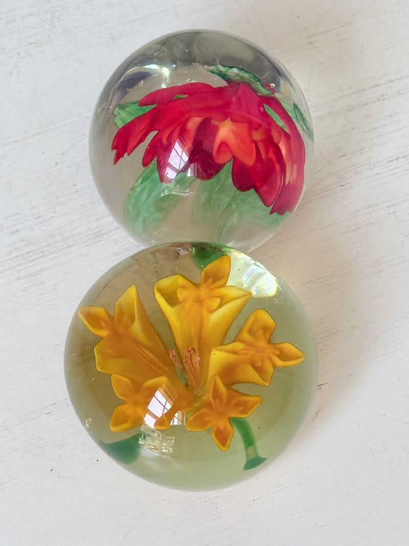 Pair of orange and yellow flower glass paperweights with green leaves, photographed on a white surface
