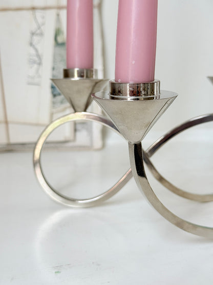 Silver tone five light candelabra with a sculptural looped base holding pink candles, photographed on a white surface