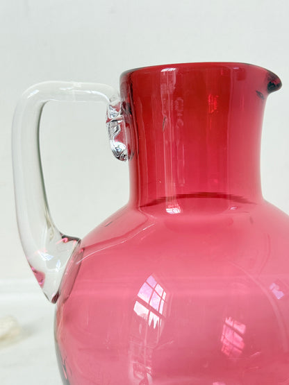 Vintage cranberry glass jug with clear handle photographed on a white surface, bulbous pink body with flared pouring rim