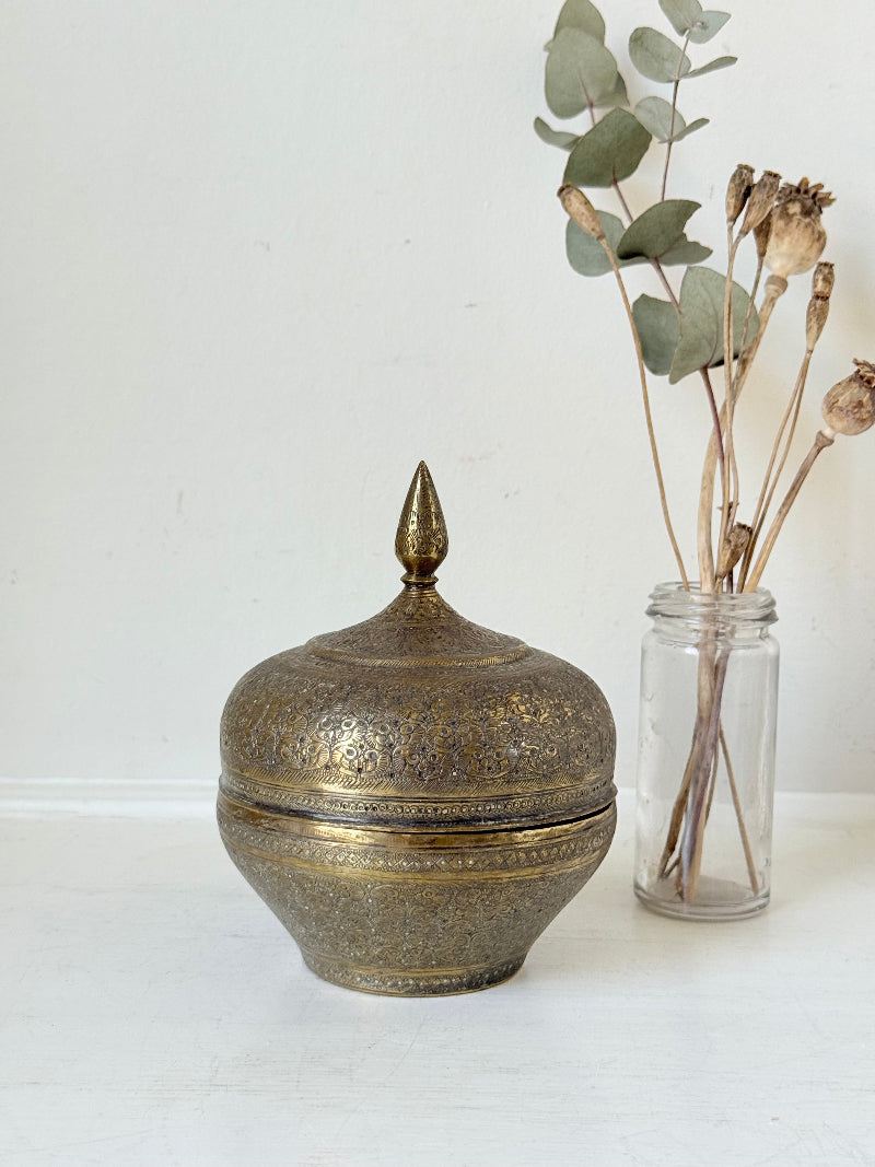 Indian Benares brass lidded jar with pointed finial and darkened interior, photographed on a white surface