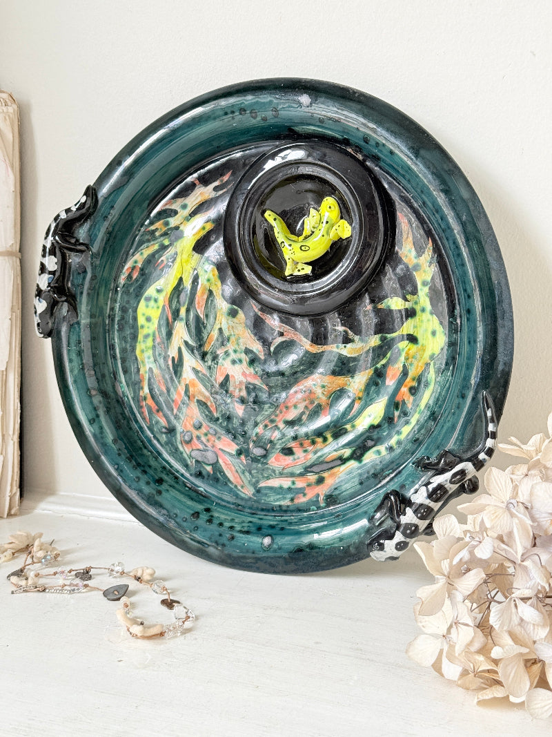 A round ceramic plate with a glossy dark green glaze, featuring a central black circular inset with a bright yellow fish, surrounded by painted orange and yellow fish motifs. The plate has black and white patterned handles on both sides and is styled on a white surface with dried flowers and a necklace.