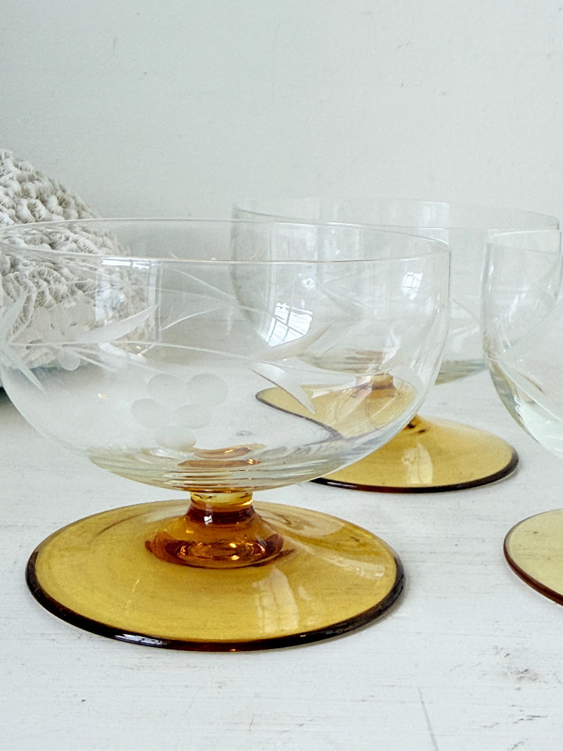 A clear glass dessert bowl with an amber-yellow circular base, shown on a white tabletop with matching glasses in the background.