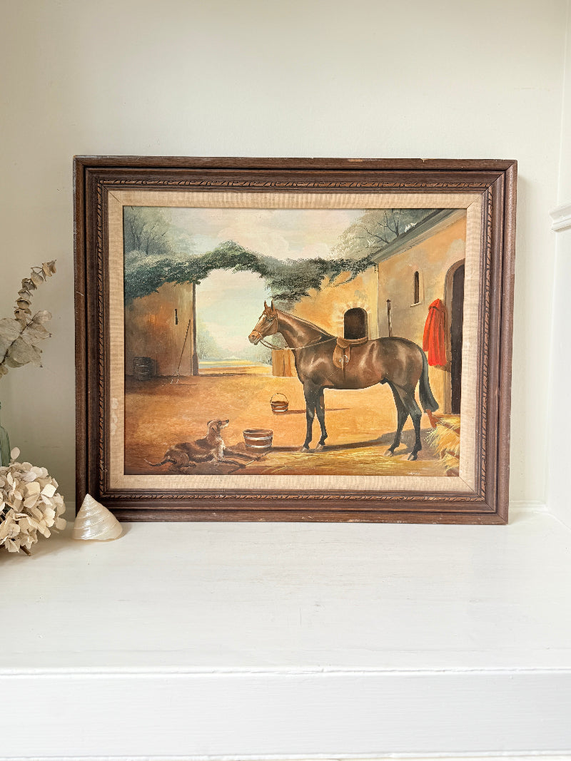 A framed vintage oil painting depicting a brown horse in a stable courtyard, displayed on a white shelf next to dried flowers.