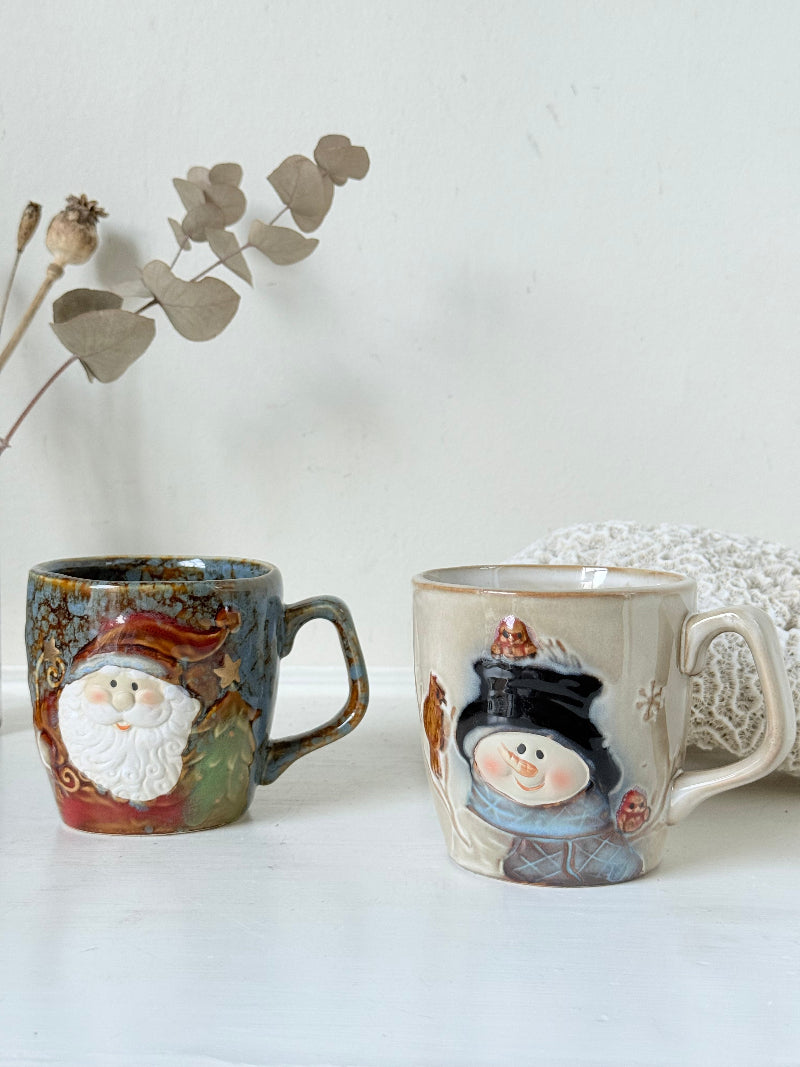 Pair of Christmas mugs with Santa and snowman designs photographed on a white surface.